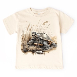 May include: Cream-colored t-shirt featuring a detailed illustration of a turtle in a natural habitat. The turtle is depicted with a patterned shell and is surrounded by tall grass and water. The design is centered on the shirt.