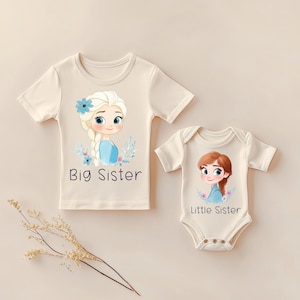 Könnte beinhalten: Zwei cremefarbene Kinderkleidungsstücke. Eines ist ein T-Shirt mit den Worten "Big Sister" und einem Cartoon-Bild eines Mädchens mit blonden Haaren. Das andere ist ein Body mit den Worten "Little Sister" und einem Cartoon-Bild eines Mädchens mit braunen Haaren.