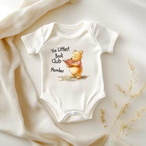 May include: White baby onesie with a graphic of Winnie the Pooh reading a book. The text "The Littlest Book Club Member" is printed above the illustration. The onesie has short sleeves and snap closures.