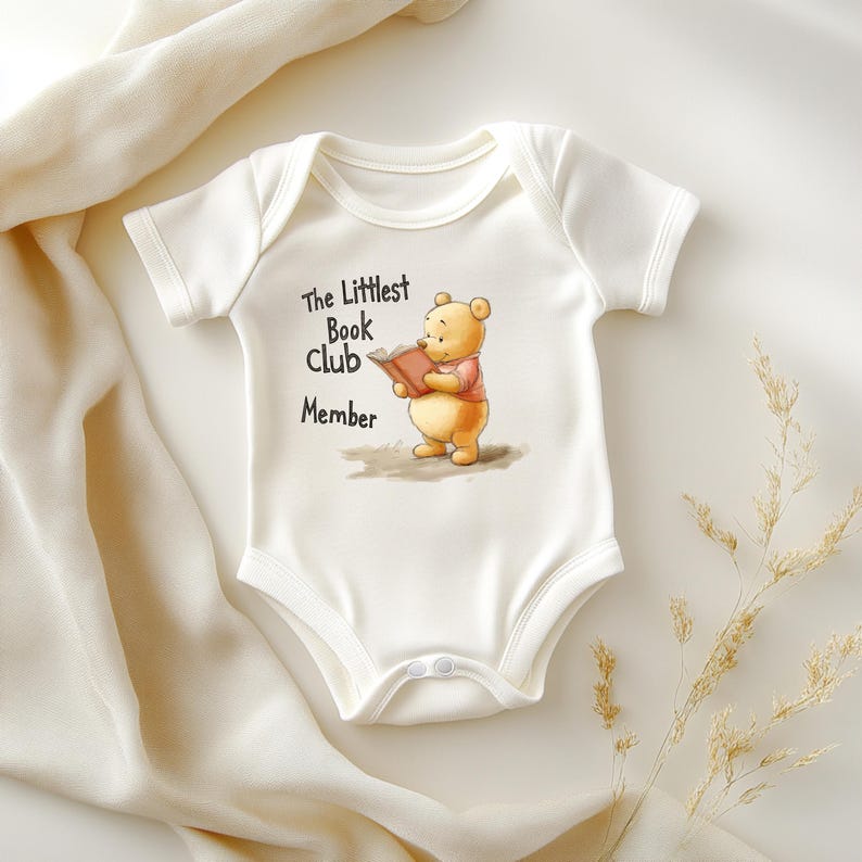 May include: White baby onesie with a graphic of Winnie the Pooh reading a book. The text "The Littlest Book Club Member" is printed above the illustration. The onesie has short sleeves and snap closures.