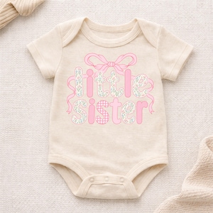May include: A light beige baby bodysuit with the words "little sister" in pink, floral, and gingham patterns. A pink bow and ribbon accent the text. The bodysuit has short sleeves and snap closures.