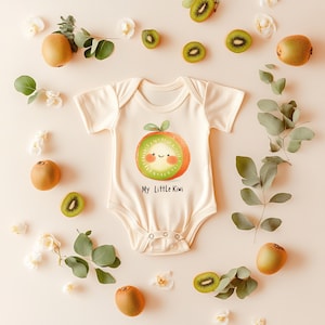 Puede incluir: Body de bebé color crema con un dibujo de kiwi sonriente y el texto "My Little Kiwi". La imagen incluye kiwis enteros, rodajas de kiwi y hojas, dispuestas sobre un fondo beige claro.