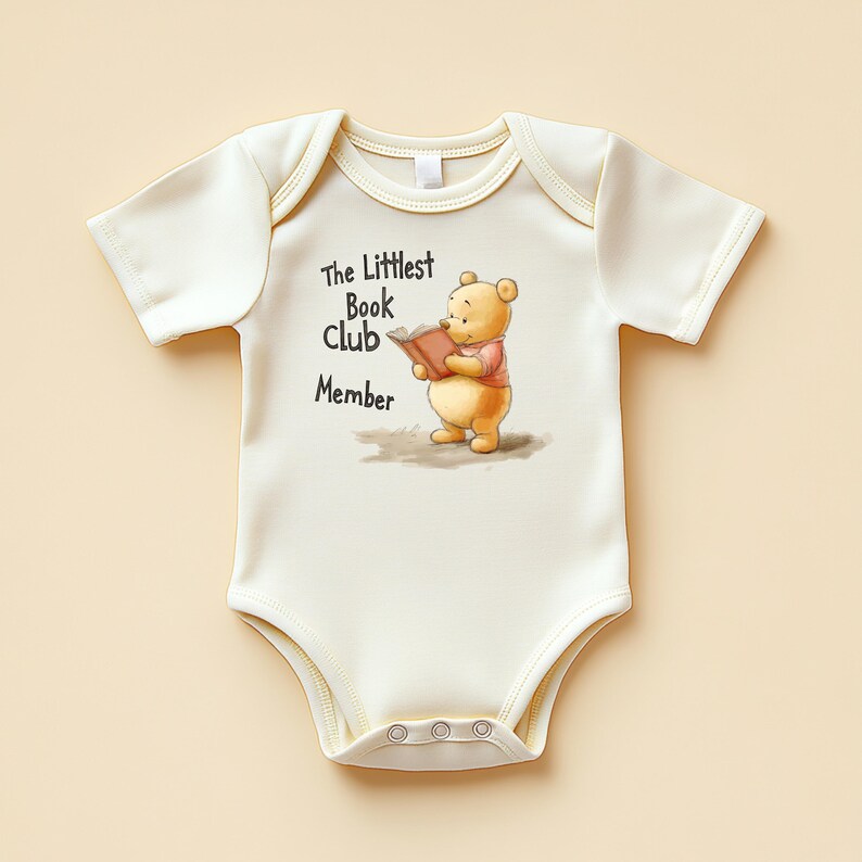 May include: A cream-colored baby onesie with short sleeves and snap closures. The onesie features a graphic of Winnie the Pooh reading a book, wearing a pink shirt. Text above the character reads 'The Littlest Book Club Member' in black.