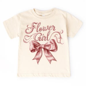 Puede incluir: Camiseta color crema con las palabras "Flower Girl" en una escritura decorativa sobre un gran lazo rosa. El lazo tiene sombreado detallado, dándole una apariencia realista. La camiseta está hecha de un material suave.