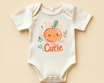 May include: A cream-coloured baby onesie featuring a cartoon orange with a smiling face and a green leaf. The text "Little Cutie" is written below the orange in orange and red.