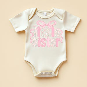 May include: Cream-colored baby bodysuit with the words "Little Sister" in pink. The word "Little" is decorated with a floral pattern, and "Sister" has a pink gingham pattern. A pink bow and ribbons accent the text.