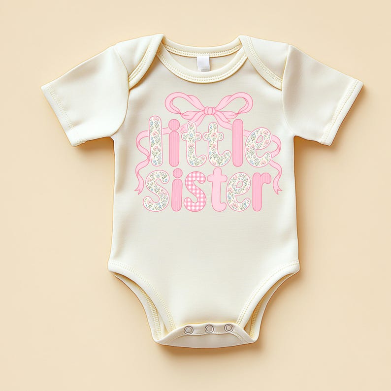 May include: Cream-colored baby bodysuit with the words "Little Sister" in pink. The word "Little" is decorated with a floral pattern, and "Sister" has a pink gingham pattern. A pink bow and ribbons accent the text.