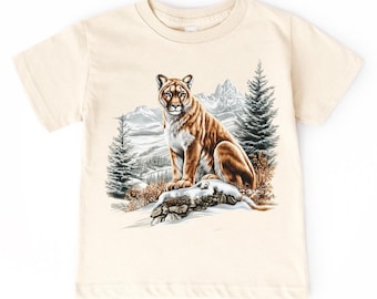 May include: A cream-colored t-shirt featuring a cougar illustration. The cougar is depicted sitting on a snow-covered rock with a mountain and pine tree backdrop. The cougar has brown fur and is facing forward.