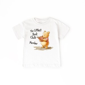 May include: White t-shirt featuring a cartoon bear reading a book, with the text "The Littlest Book Club Member" in black. The bear is wearing a pink shirt. A children's apparel item.