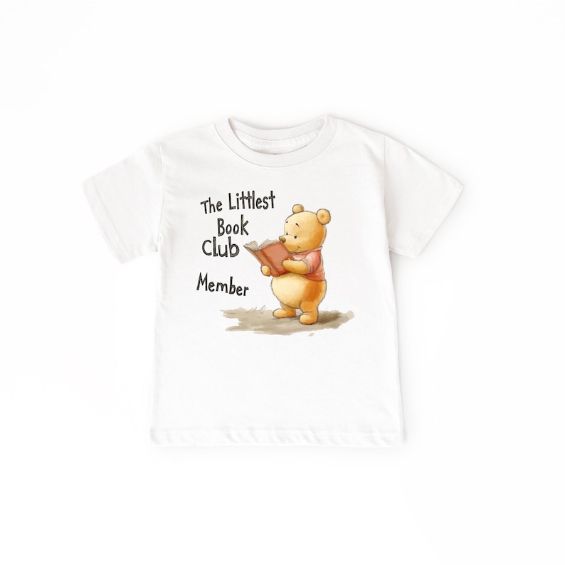 May include: White t-shirt featuring a cartoon bear reading a book, with the text "The Littlest Book Club Member" in black. The bear is wearing a pink shirt. A children's apparel item.
