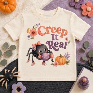 May include: A cream-colored short-sleeve t-shirt featuring a graphic of a fuzzy black spider wearing a bow and sneakers, holding a purple polka-dot candy. The text reads Creep It Real in orange and purple, surrounded by orange flowers and a pumpkin.