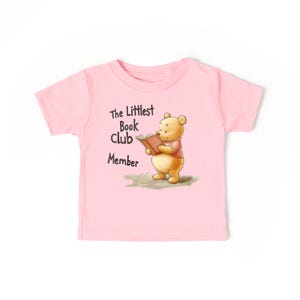 May include: A light pink toddler t-shirt featuring a cartoon illustration of a bear reading a book. The text "The Littlest Book Club Member" is printed above the bear. The bear is wearing a pink shirt and holding a brown book.