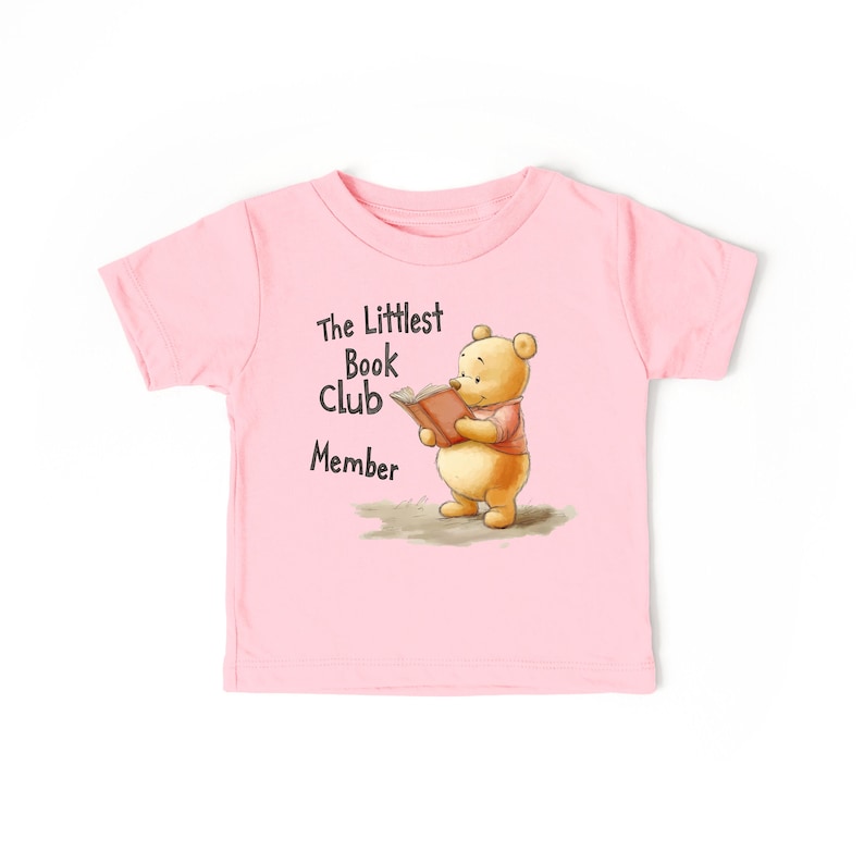 May include: A light pink toddler t-shirt featuring a cartoon illustration of a bear reading a book. The text "The Littlest Book Club Member" is printed above the bear. The bear is wearing a pink shirt and holding a brown book.