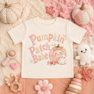 May include: A cream-colored short-sleeve t-shirt featuring the text "Pumpkin Patch Babe" in pink and orange tones, accompanied by a graphic of a smiling pumpkin and a small ghost, surrounded by decorative floral and vine illustrations.