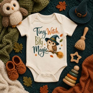 May include: A cream-colored short-sleeve baby bodysuit featuring a graphic of an owl wearing a witch hat on a broomstick. The text reads Tiny Witch, BIG Magic. The design includes small orange and green leaves and stars surrounding the central owl illustration.