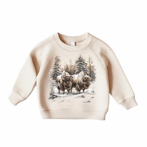 May include: Cream-colored sweatshirt featuring a detailed illustration of two bison in a snowy forest scene. The graphic is in shades of brown and gray. The sweatshirt has a crew neck and long sleeves.