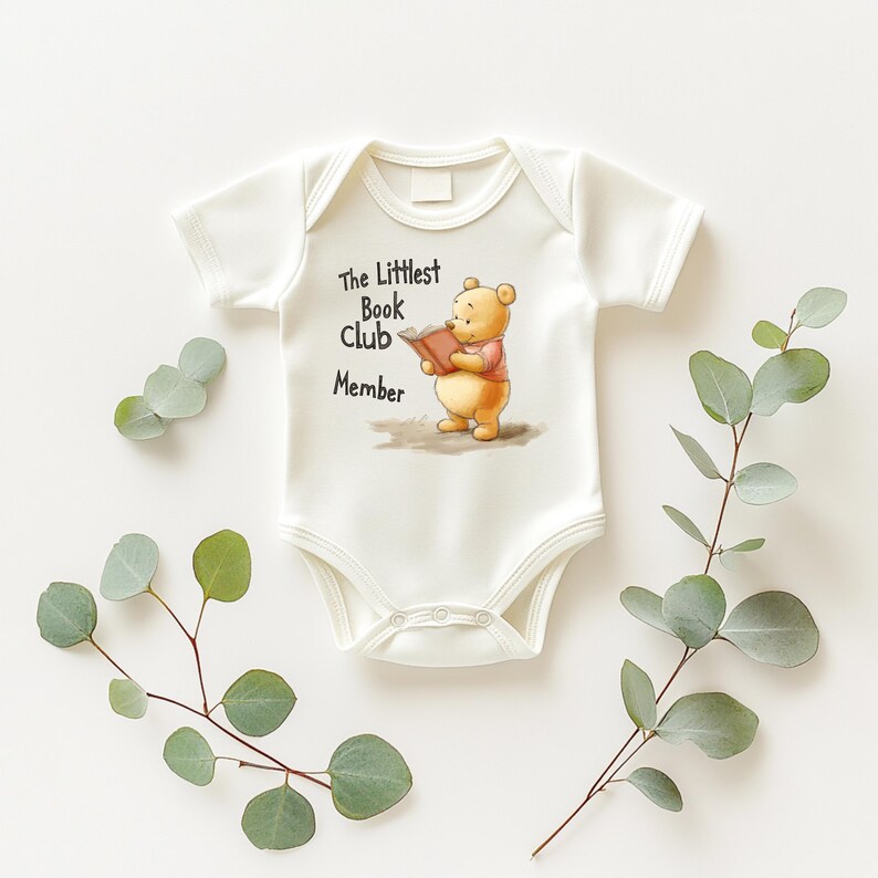 May include: White baby onesie with short sleeves. The onesie features a graphic of a cartoon bear reading a book, with the text "The Littlest Book Club Member". The onesie is surrounded by green leafy sprigs.