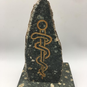 May include: A dark gray stone sculpture featuring a gold-colored caduceus symbol. The stone has a rough, natural edge and sits on a matching base. The caduceus, a symbol of medicine, is carved into the stone.