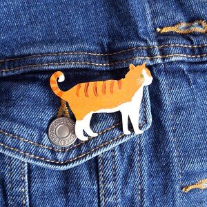 May include: A decorative brooch shaped like a standing cat with orange tabby stripes on its back and head, a white chest, white legs, and a curled tail. The cat has a small green eye and is positioned against a dark blue denim fabric background.