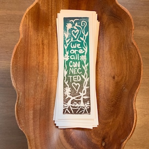 May include: A stack of bookmarks with a green and brown gradient background. The bookmarks feature a white print of a heart, vines, and the words "We are all connected".