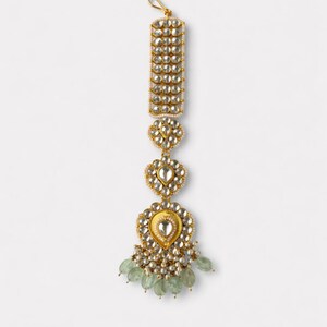 May include: A gold-toned maang tikka headpiece featuring a rectangular top section, two floral-shaped middle links, and a large teardrop-shaped base. The piece is adorned with clear stones, small white pearls, and pale green teardrop beads.
