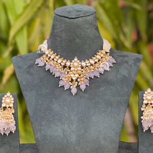 May include: A gold-toned necklace and earring set with a floral design. The necklace features a central pendant with clear stones and lavender-colored bead drops. The matching earrings have a similar design, with clear stones and lavender beads.