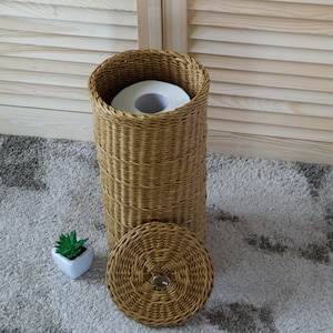 May include: A woven wicker toilet paper holder with a lid and a roll of toilet paper inside. The holder is sitting on a light gray rug.
