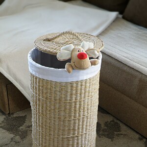 May include: A woven wicker laundry hamper with a white fabric liner and a removable lid. A plush reindeer toy is peeking out of the top of the hamper.