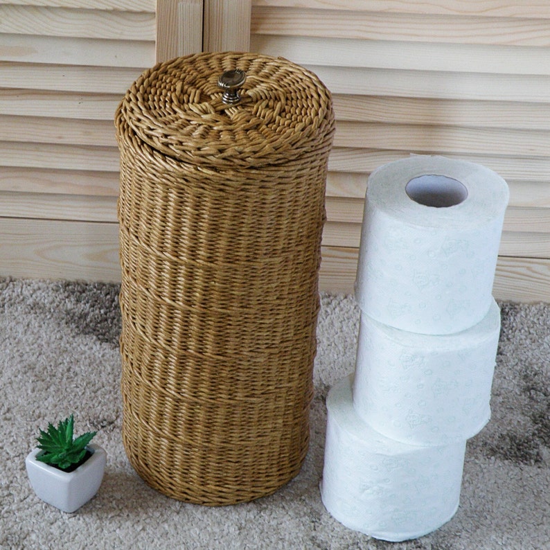 May include: A woven wicker toilet paper holder with a lid and a stack of three white toilet paper rolls.