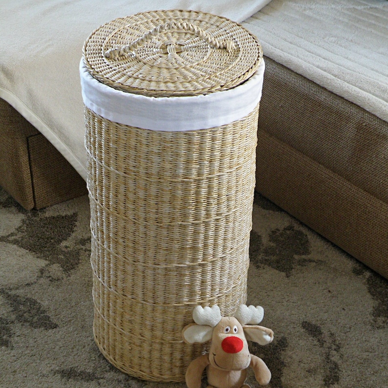 May include: A tall, cylindrical wicker laundry hamper with a white fabric liner and a woven lid. The hamper is sitting on a patterned rug with a small stuffed reindeer toy in front of it.