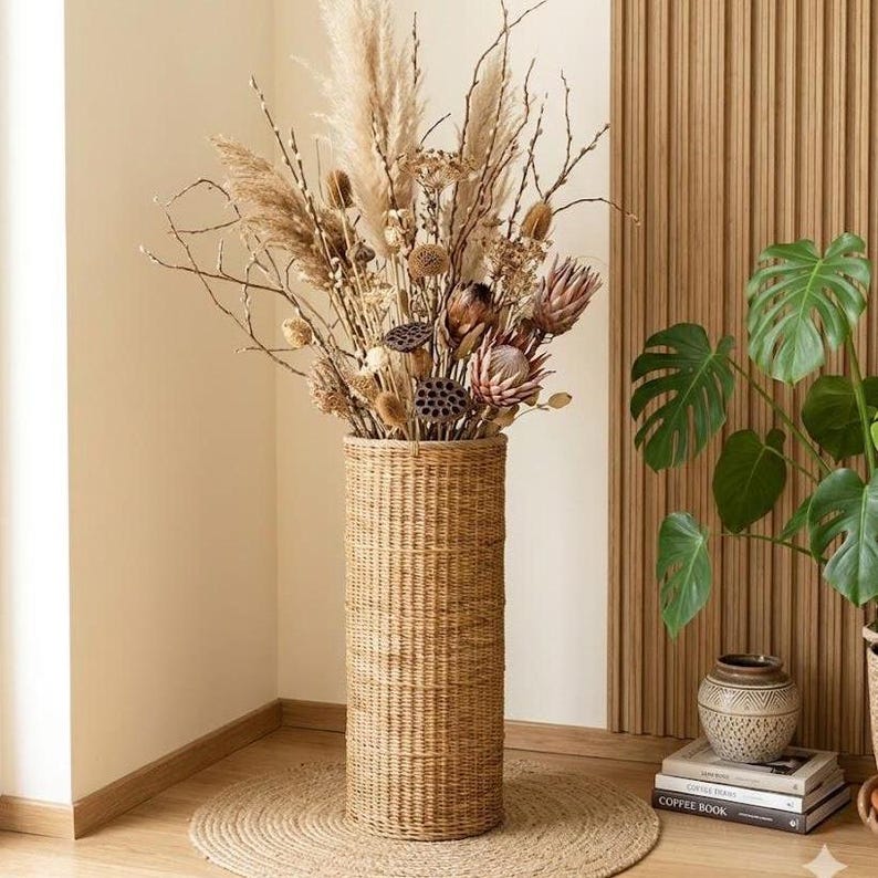 May include: A tall, cylindrical floor vase woven from light brown natural rattan. The vase is filled with a large arrangement of dried beige pampas grass, brown lotus seed pods, and textured protea flowers in shades of tan and muted pink.