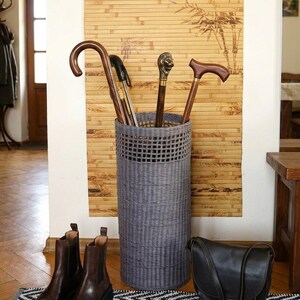 May include: A tall, cylindrical grey woven basket used as an umbrella stand, containing four walking sticks with various handles, including one with a metallic lion head topper, positioned upright in the centre of the frame.