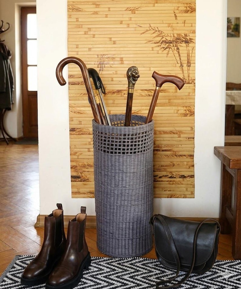 May include: A tall, cylindrical grey woven basket used as an umbrella stand, containing four walking sticks with various handles, including one with a metallic lion head topper, positioned upright in the centre of the frame.