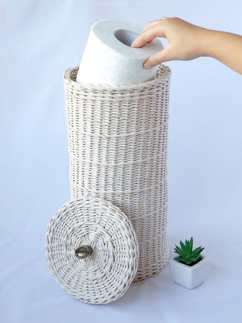 May include: White wicker toilet paper holder with a lid and a brass knob. The holder is filled with a roll of toilet paper.
