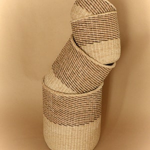 May include: A stack of four cylindrical woven baskets made from natural light beige and dark brown fibers. The baskets are arranged in a vertical, slightly tilted column, each featuring a distinct horizontal band of dark brown woven pattern.