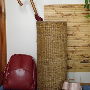 May include: A tall cylindrical woven wicker umbrella stand with a natural tan finish. Inside the stand is a dark red umbrella with a curved wooden handle. Beside the stand is a dark red leather backpack and a pair of light pink heeled sandals.