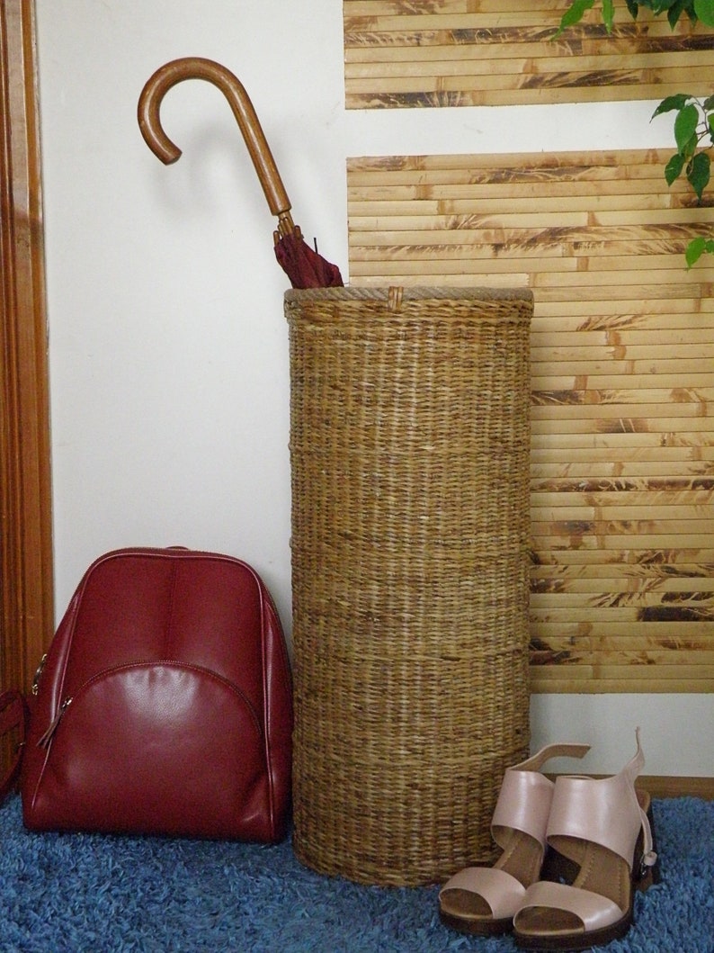 May include: A tall cylindrical woven wicker umbrella stand with a natural tan finish. Inside the stand is a dark red umbrella with a curved wooden handle. Beside the stand is a dark red leather backpack and a pair of light pink heeled sandals.