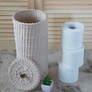 Könnte beinhalten: Ein weißer Toilettenpapierhalter aus Weidengeflecht mit Deckel und einer kleinen Topfpflanze. Der Halter steht auf einem beigefarbenen Teppich, neben ihm liegen drei Rollen Toilettenpapier übereinander.