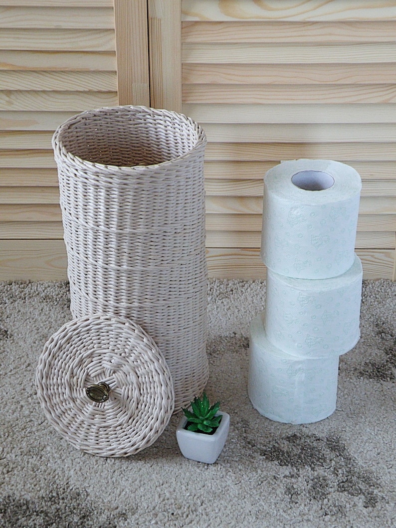 May include: A white wicker toilet paper holder with a lid and a small potted succulent plant. The holder is sitting on a beige carpet with three rolls of toilet paper stacked next to it.
