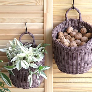 May include: Two woven brown baskets hanging on a wooden shutter. One basket holds green and white leafy plants, and the other is filled with walnuts. The baskets have a handle for hanging and are a decorative storage solution.