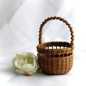 May include: A small, woven brown basket with a handle, next to a large white flower with green leaves. The basket is round with a woven handle. The flower has multiple petals and is slightly open.