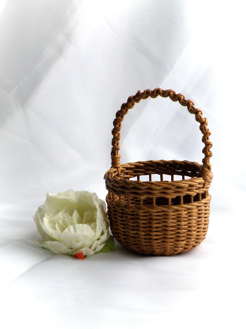 May include: A small, woven brown basket with a handle, next to a large white flower with green leaves. The basket is round with a woven handle. The flower has multiple petals and is slightly open.