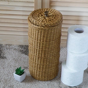 May include: A woven wicker toilet paper holder with a brown finish and a metal top. Three rolls of white toilet paper are stacked next to the holder.