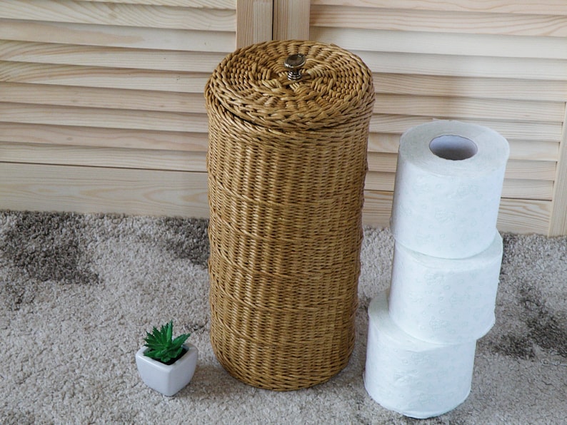 May include: A woven wicker toilet paper holder with a brown finish and a metal top. Three rolls of white toilet paper are stacked next to the holder.