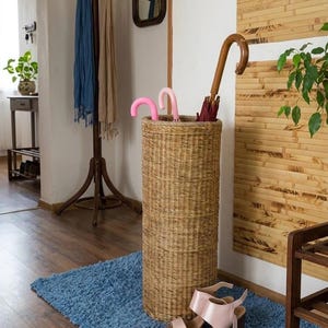 May include: A tall, cylindrical, woven wicker umbrella stand in a natural tan colour. The stand holds two small pink umbrellas with curved handles and one larger umbrella with a dark red canopy and a curved wooden handle, placed on a blue rug.