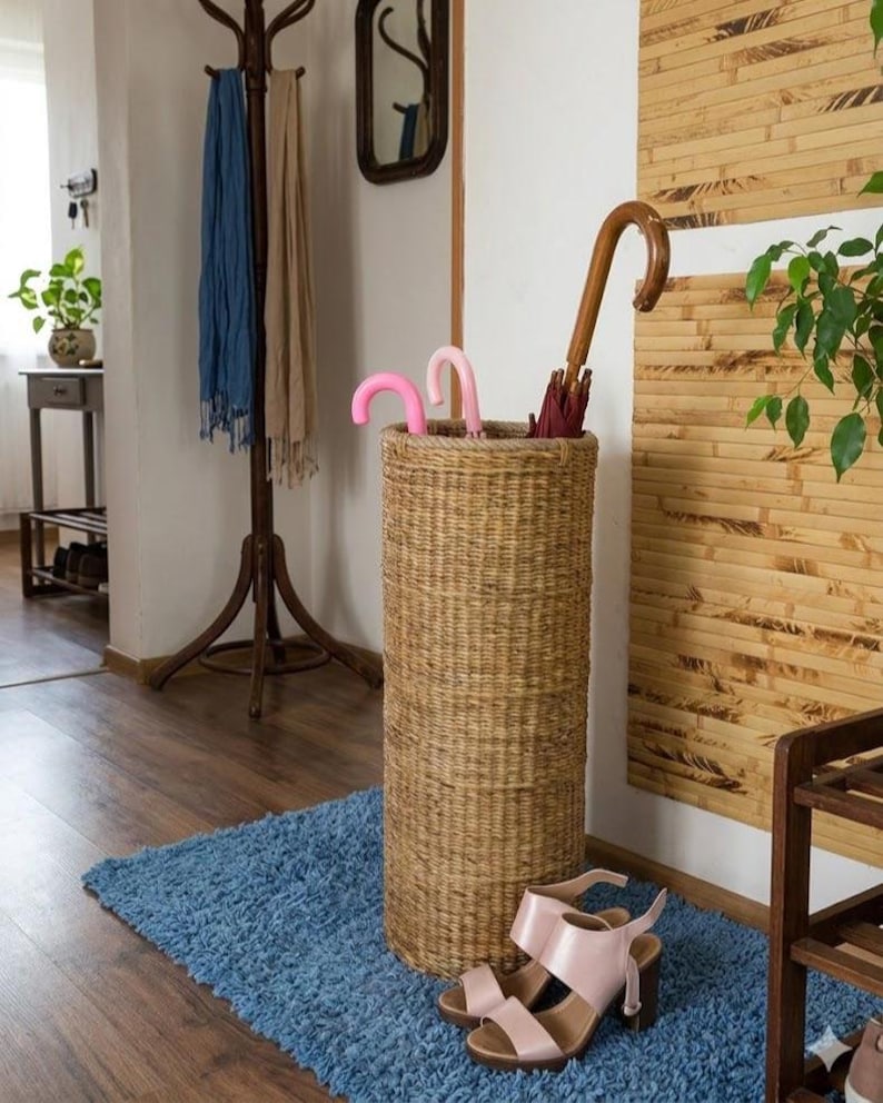 May include: A tall, cylindrical, woven wicker umbrella stand in a natural tan colour. The stand holds two small pink umbrellas with curved handles and one larger umbrella with a dark red canopy and a curved wooden handle, placed on a blue rug.