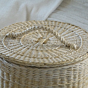 May include: A white wicker basket with a lid and a woven handle. The basket is round and has a natural, rustic look.