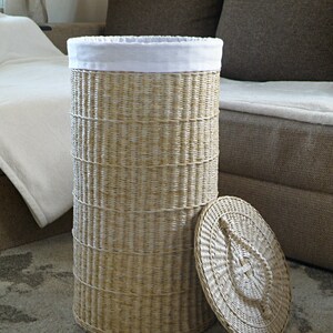 May include: A woven wicker laundry basket with a white fabric liner and a matching lid. The basket is sitting on a rug.