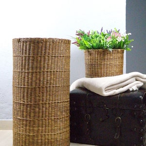 Op de afbeelding: Twee bruine rieten manden, een groter dan de andere, met een wit deken en een zwarte houten kist op de achtergrond. De grotere mand is leeg, terwijl de kleinere mand een plant in een pot met groene bladeren en roze en paarse bloemen bevat.