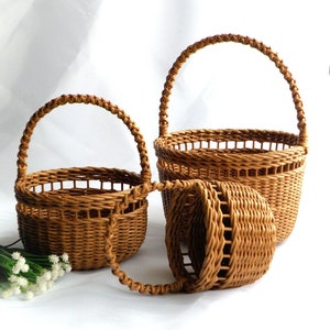 May include: Three brown woven baskets of different sizes, each with an arched handle. The baskets are made from a light brown material and feature a woven pattern. The handles are also woven and curved. They are arranged on a white surface.